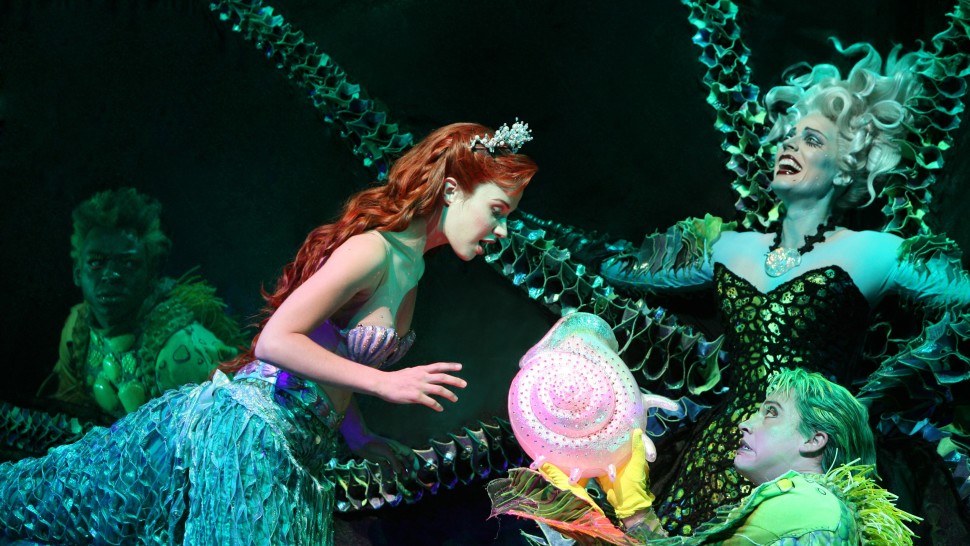 Scene from The Little Mermaid on Broadway featuring red-headed Ariel and Ursula the sea witch. Ariel is singing into a shell.